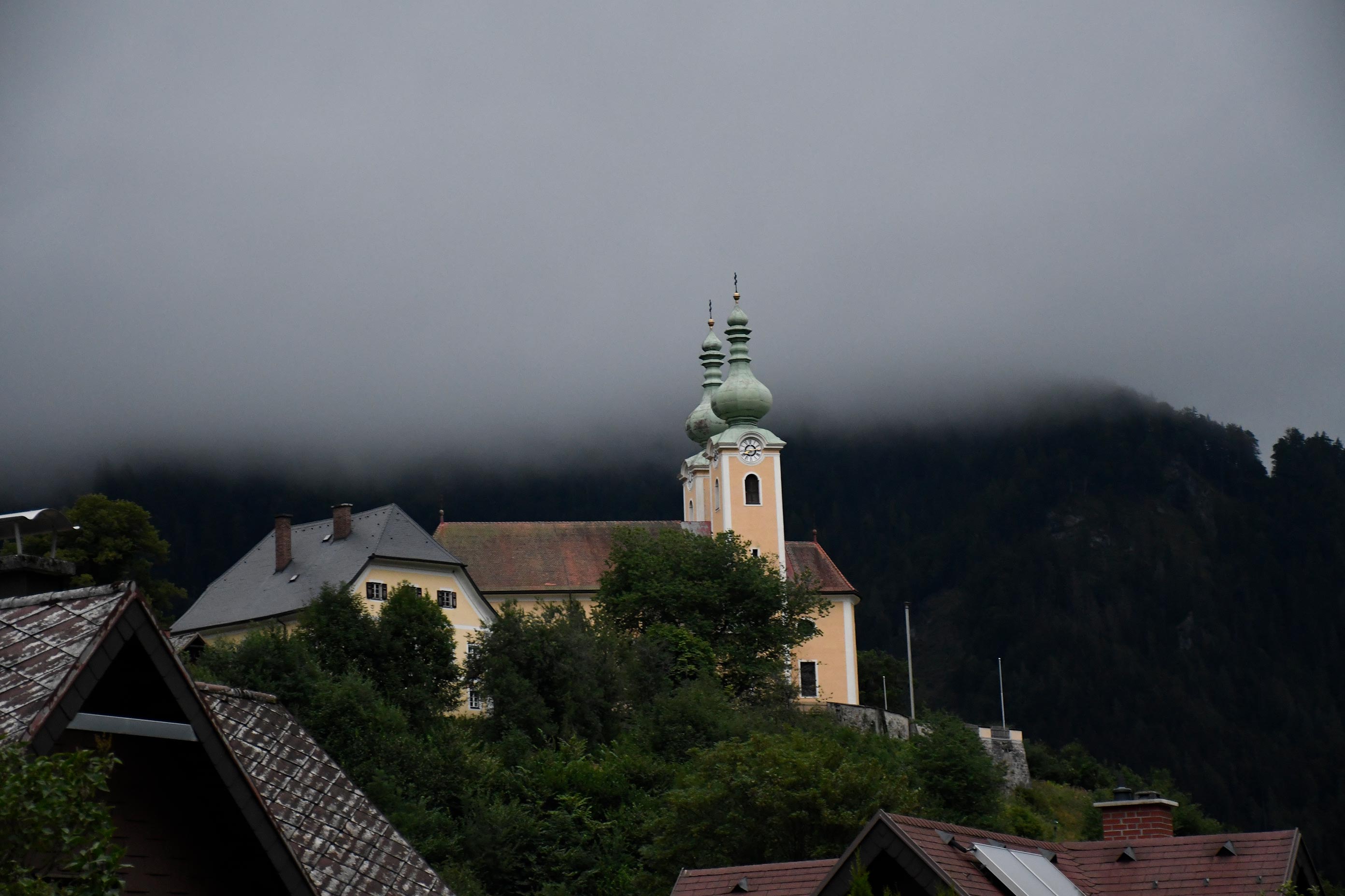 Ein Bild einer Kirche auf einem Hügel, umringt von Bäumen. Im Hintergrund ist ein vernebelter Berg.